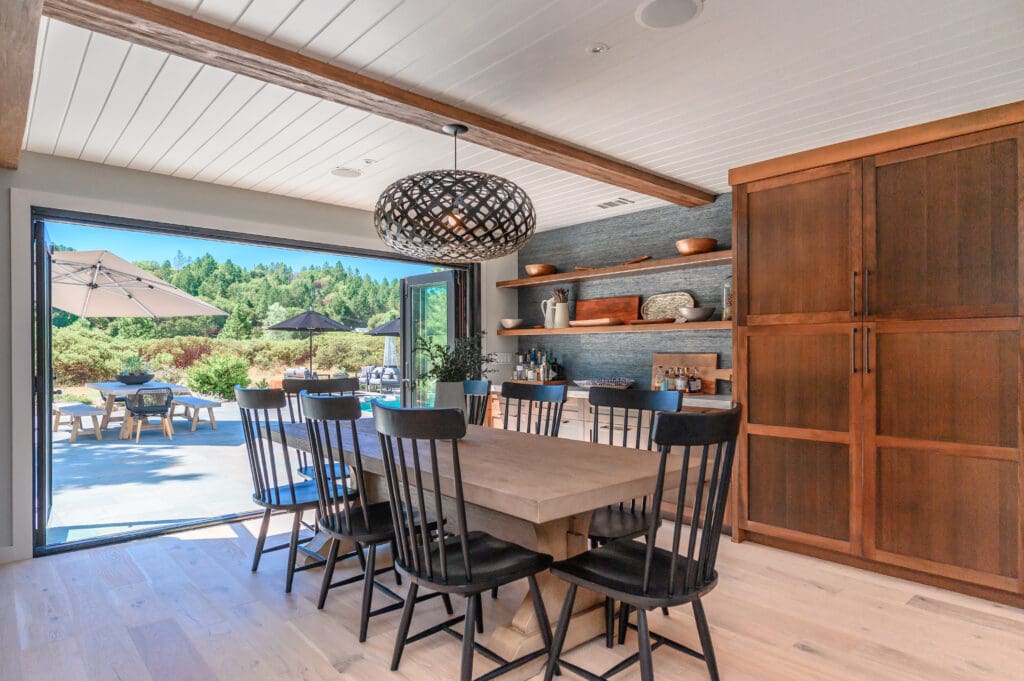 Dining room with large wood table and black spindle chairs opening to an outdoor patio through folding glass doors.