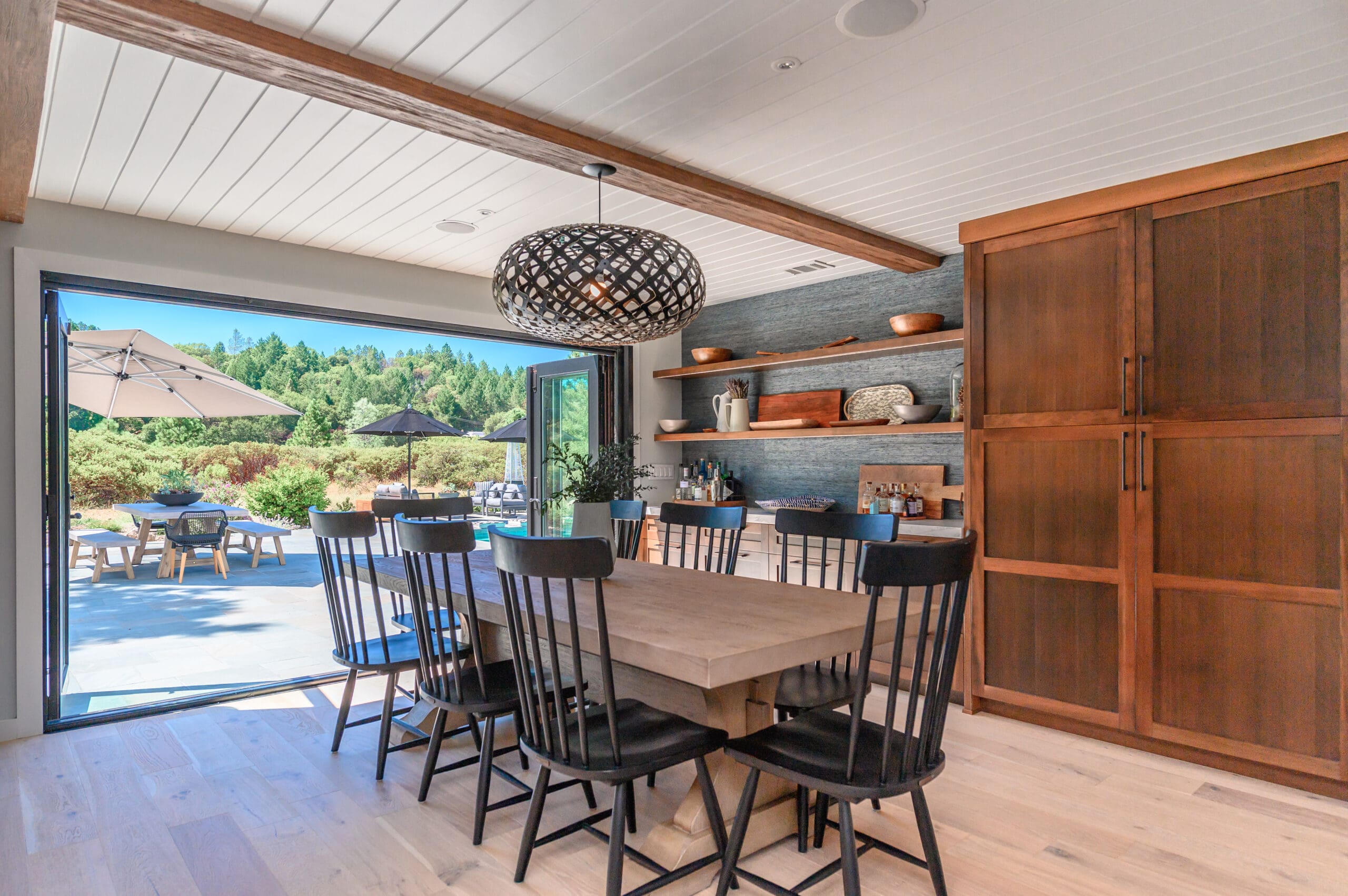 Dining room with large wood table and black spindle chairs opening to an outdoor patio through folding glass doors.